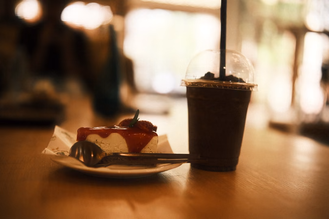 Slice of cheesecake with red berry topping and a fork beside an iced dark drink in a domed cup on a wooden table