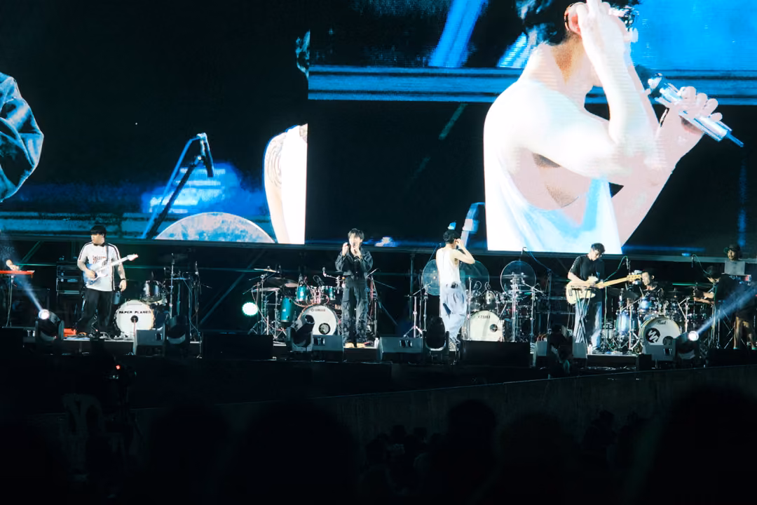 Band on stage below a screen showing a close-up of the singer lifting a microphone, blue light on screen.