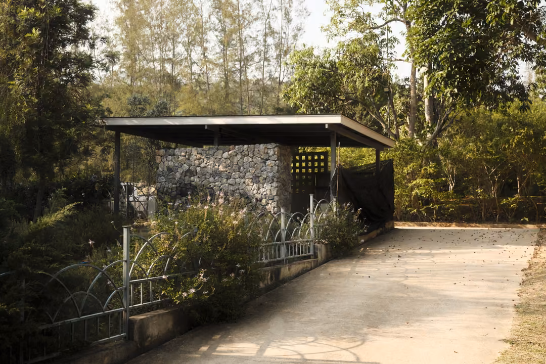 A small open shelter with a stone wall at the end of a concrete drive, trees around it