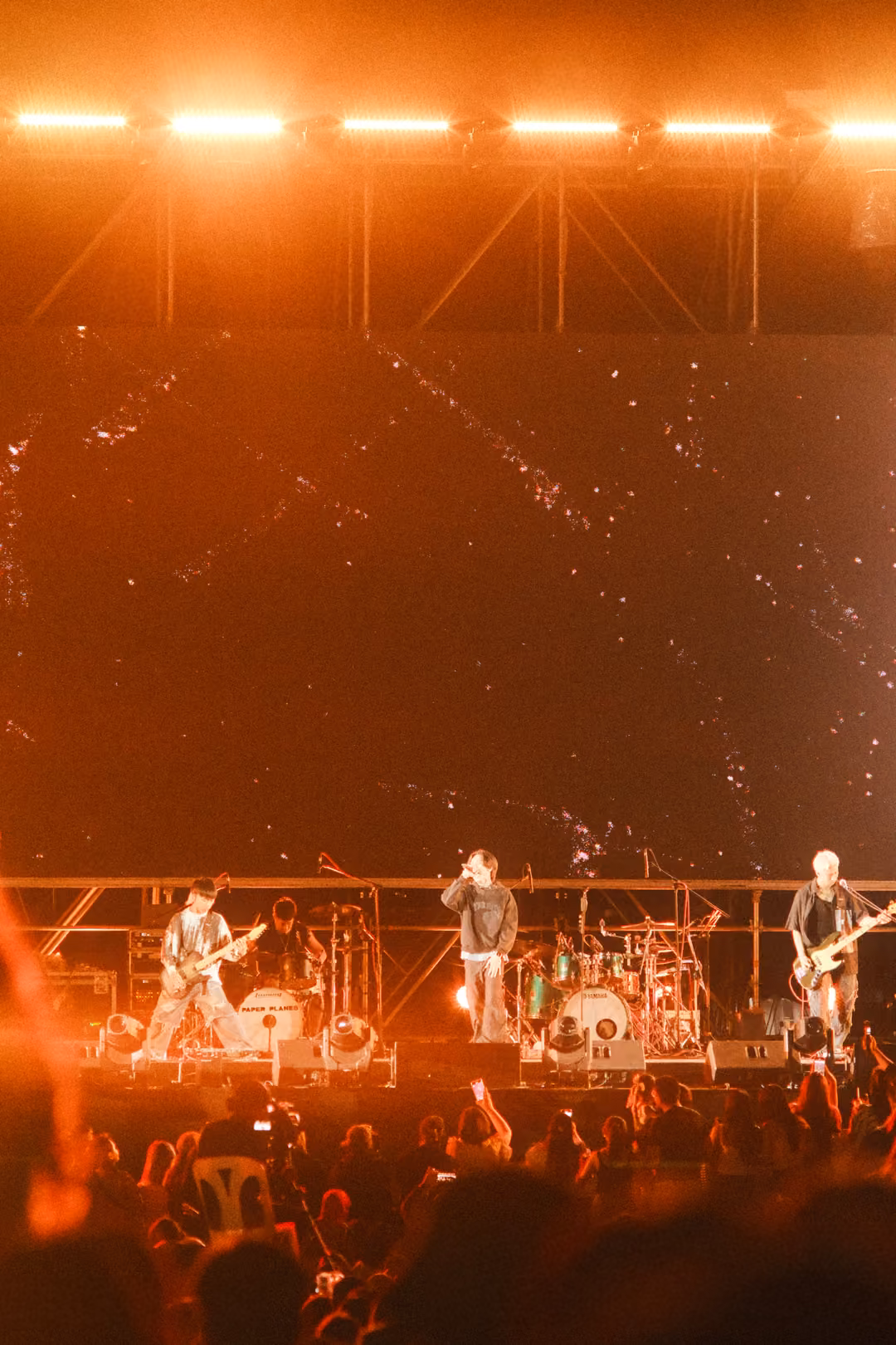 Vertical view of a stage in warm orange light, guitarists and singer performing, crowd with raised hands below.