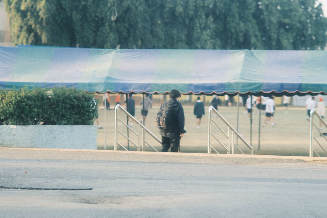A figure with a backpack walks between railings under a striped green and blue tent, students on a field beyond.