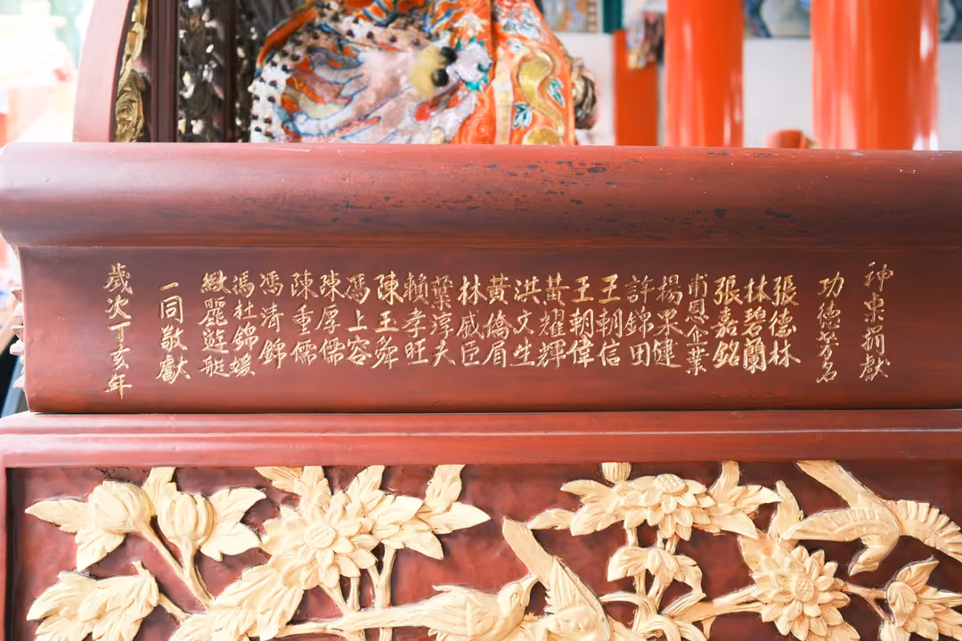 Red lacquered panel inscribed with gold names in columns above a gilded carving of flowers and birds