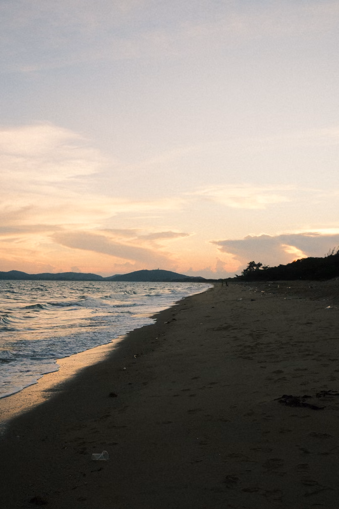 Vertical view of a long empty beach at sunset with gentle waves, distant hills and a pastel sky