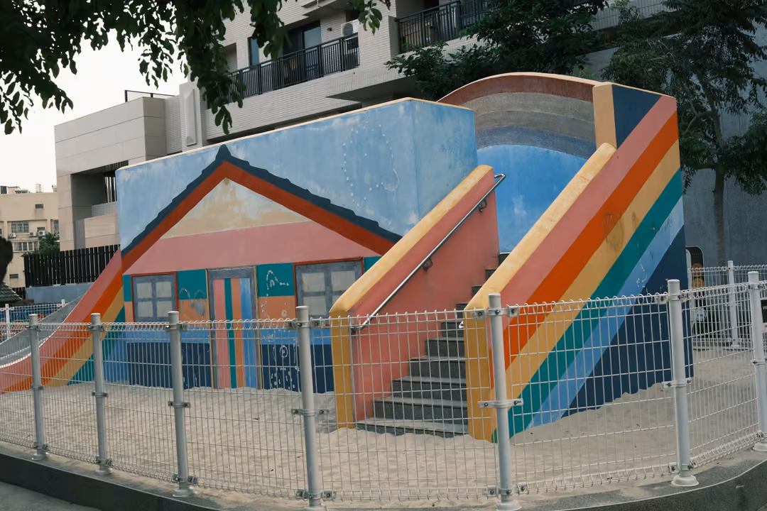 Playground structure painted with rainbow stripes and a house shape, behind a metal fence