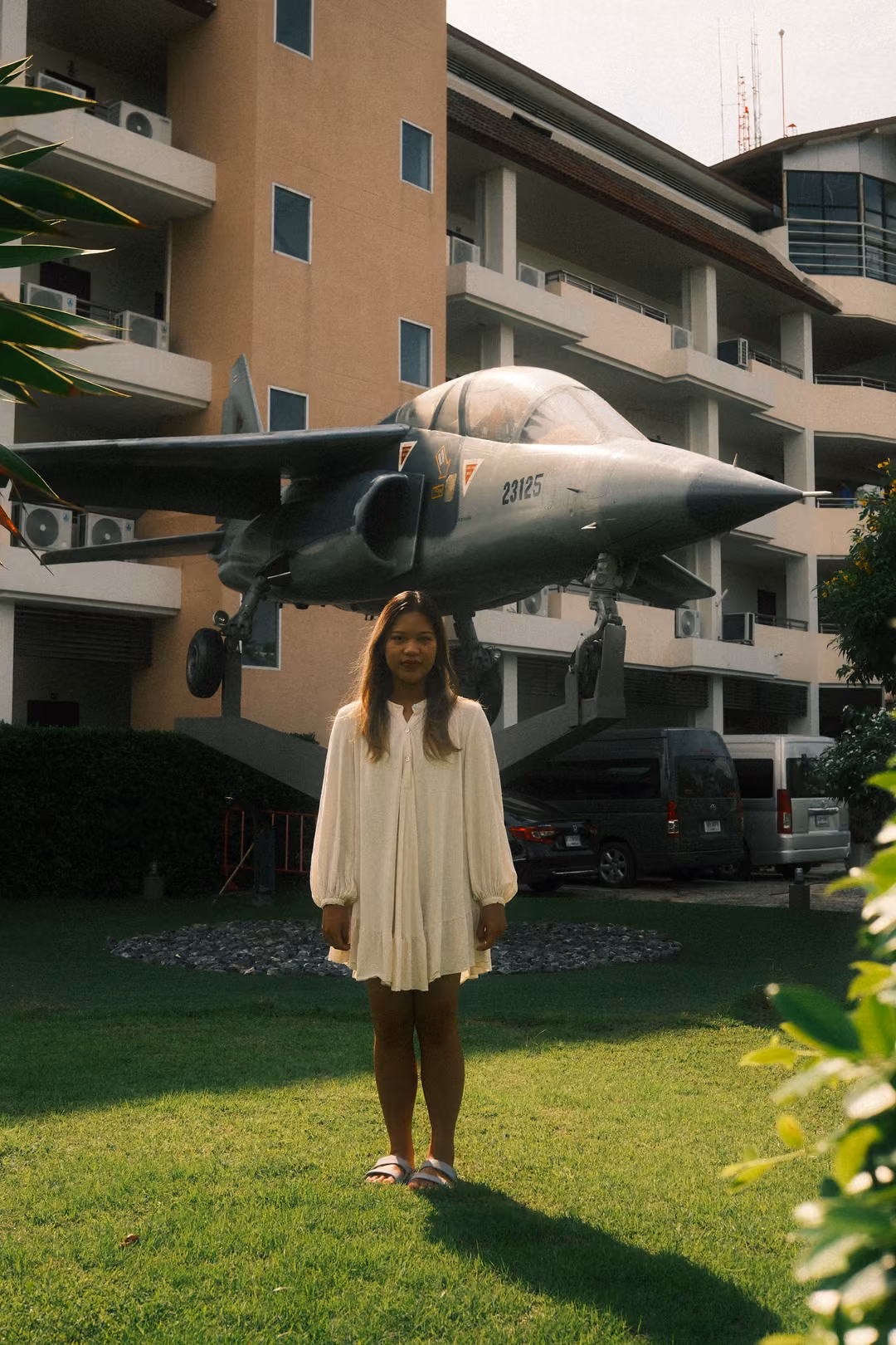Woman in a white dress standing on a lawn beneath a grey jet fighter mounted on a pedestal in front of an apartment block
