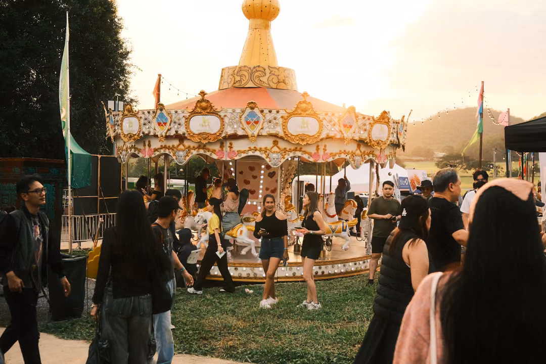 Crowd walking past a gold-and-white carousel on a grassy field, backlit by a hazy evening sun.