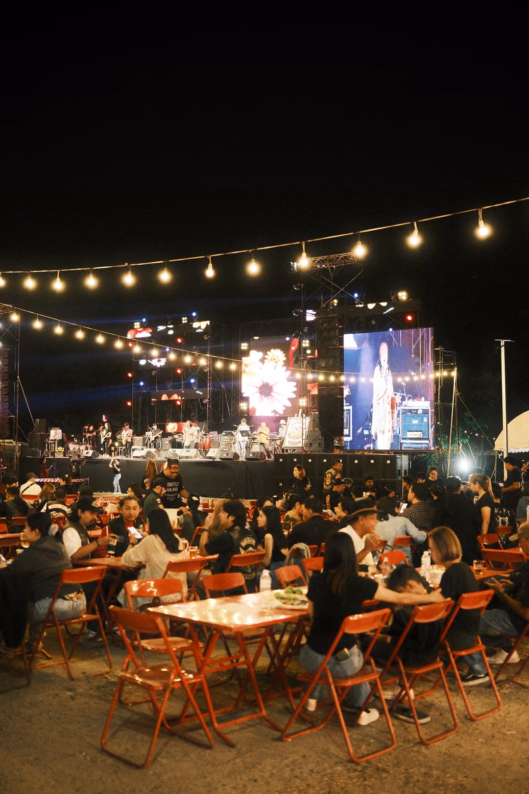 Night festival seating area with red tables and chairs, string lights and a glowing stage screen
