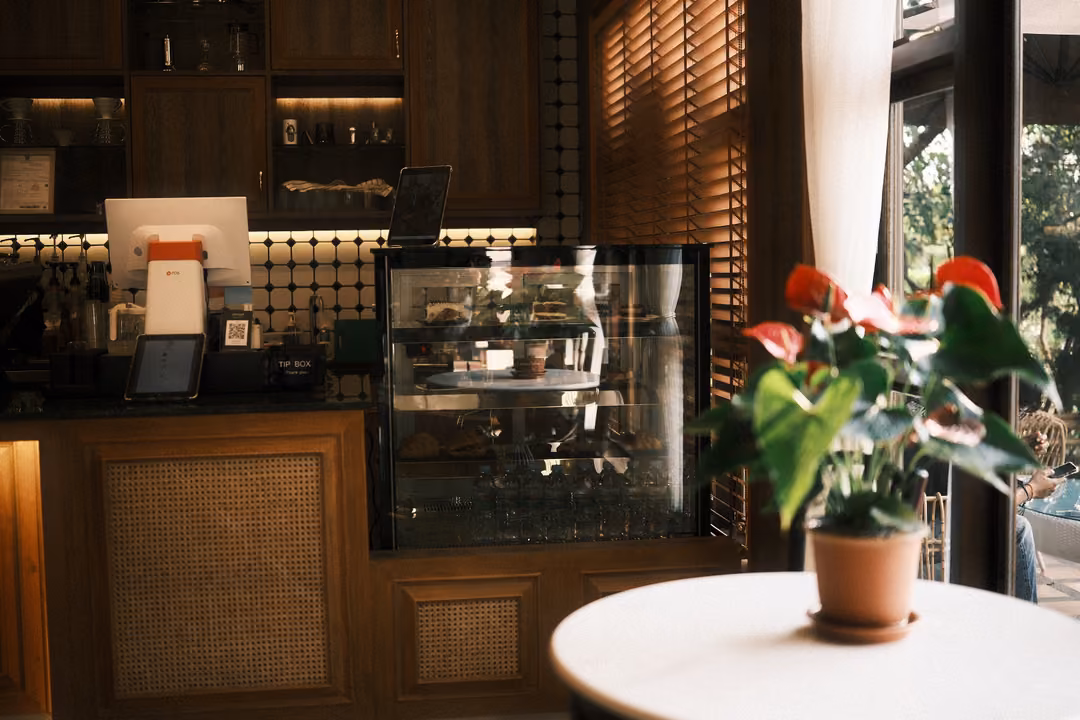 Potted red anthurium on a round white table in front of a café counter, pastry case and wooden blinds