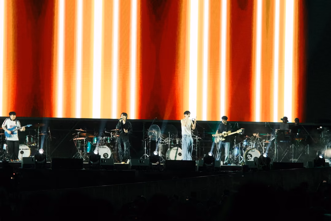 Band performing in front of a red and orange screen of narrow vertical light stripes, white beams fanning upward.