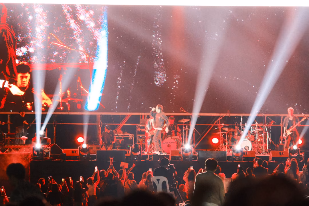 Stage with white light beams fanning upward through red haze, band performing, crowd with raised phones in front.