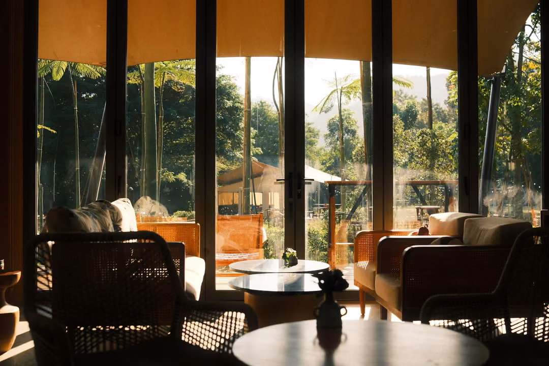 Dim lounge with wicker armchairs and round tables silhouetted against tall bright windows and garden