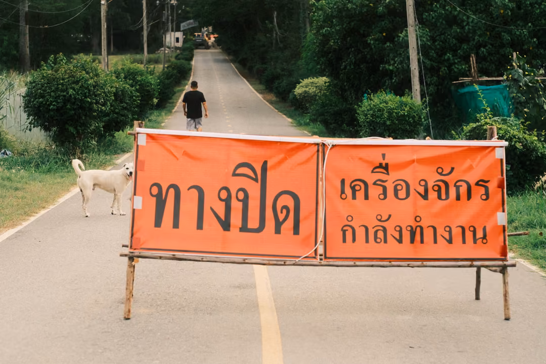 An orange Thai road-closed banner on a wooden stand across a road, a white dog beside it and a figure walking beyond.