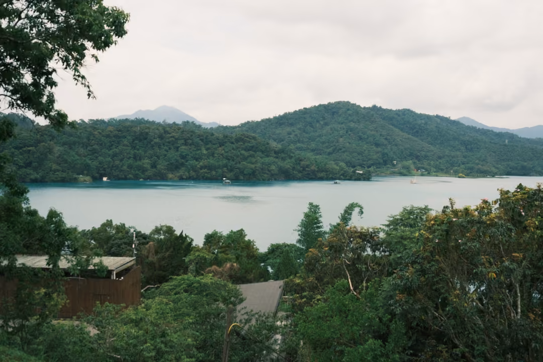 Elevated view over treetops and a rooftop to a wide lake and layered green mountains under cloud