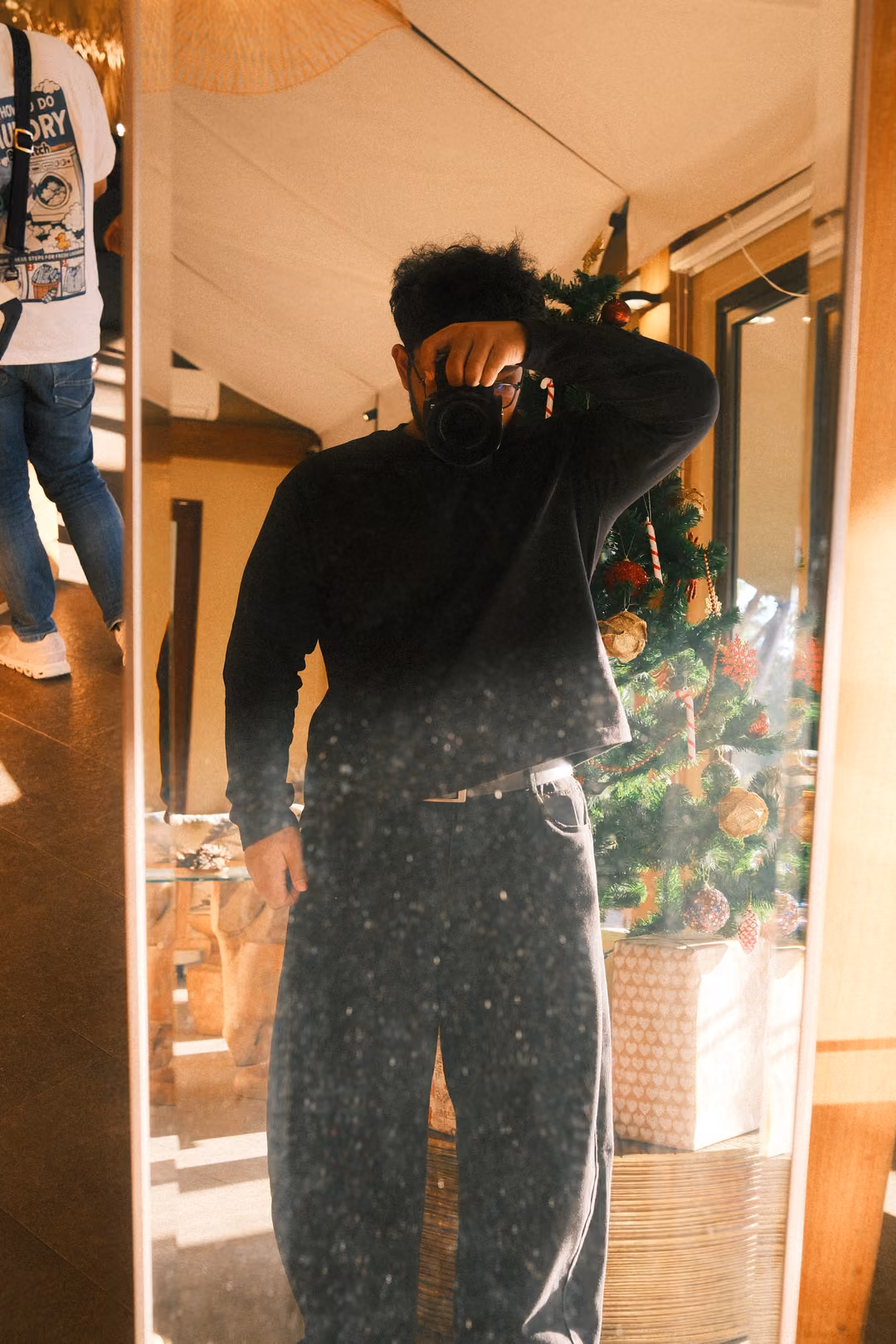 Person in black reflected in a tall standing mirror, one hand raised to the face, Christmas tree behind