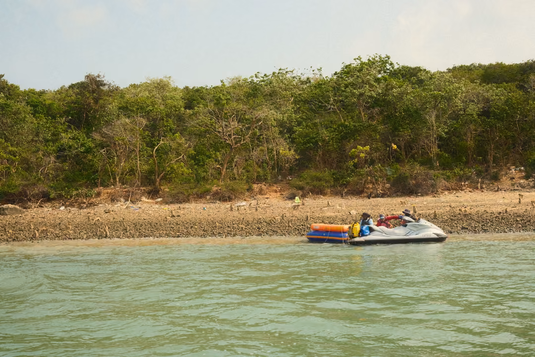 A jet ski towing a colourful inflatable banana pulls up to a rocky shore below green trees.