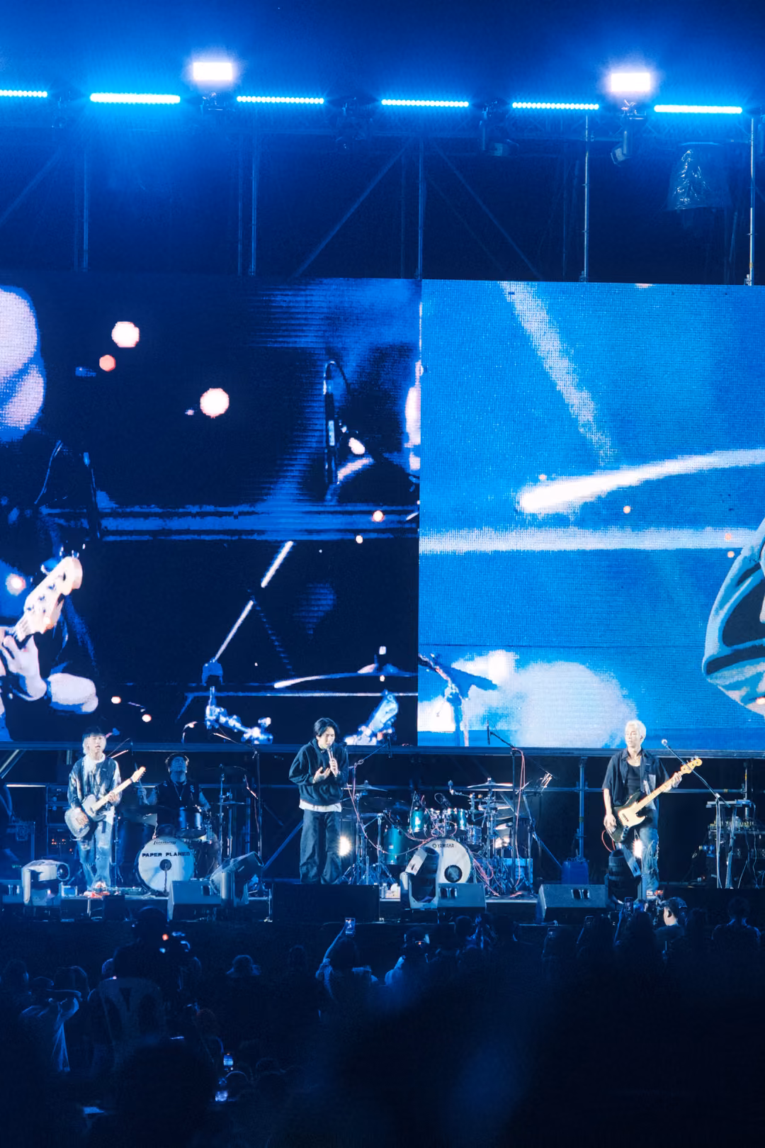 Vertical view of a screen with blue streaked panels above three musicians performing, blue lamps on the truss.