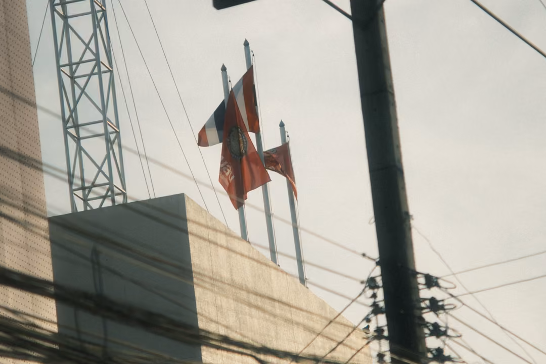 Flags on tall poles atop a building beside a steel lattice tower, with power lines and a utility pole in front.