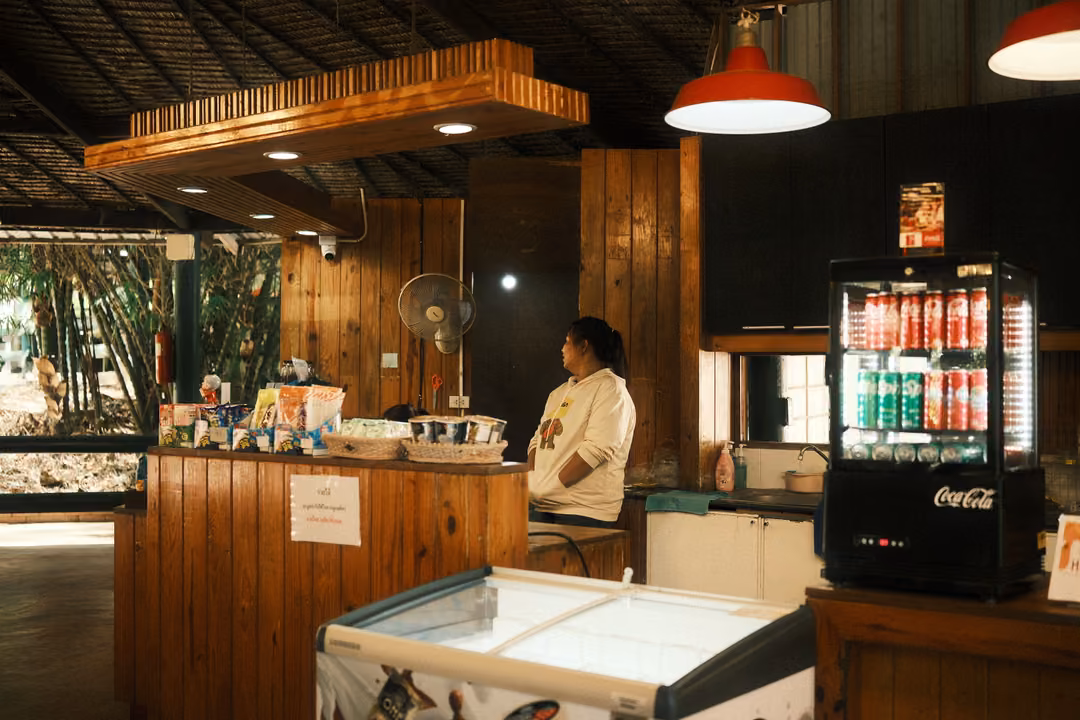 Wooden snack kiosk with red pendant lamps, a drinks fridge and a person seated at the counter