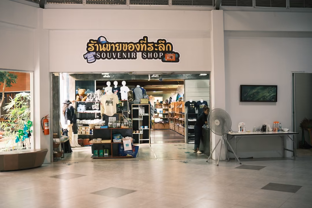 A shop entrance under a sign reading Souvenir Shop in Thai and English, T-shirts and shelves inside