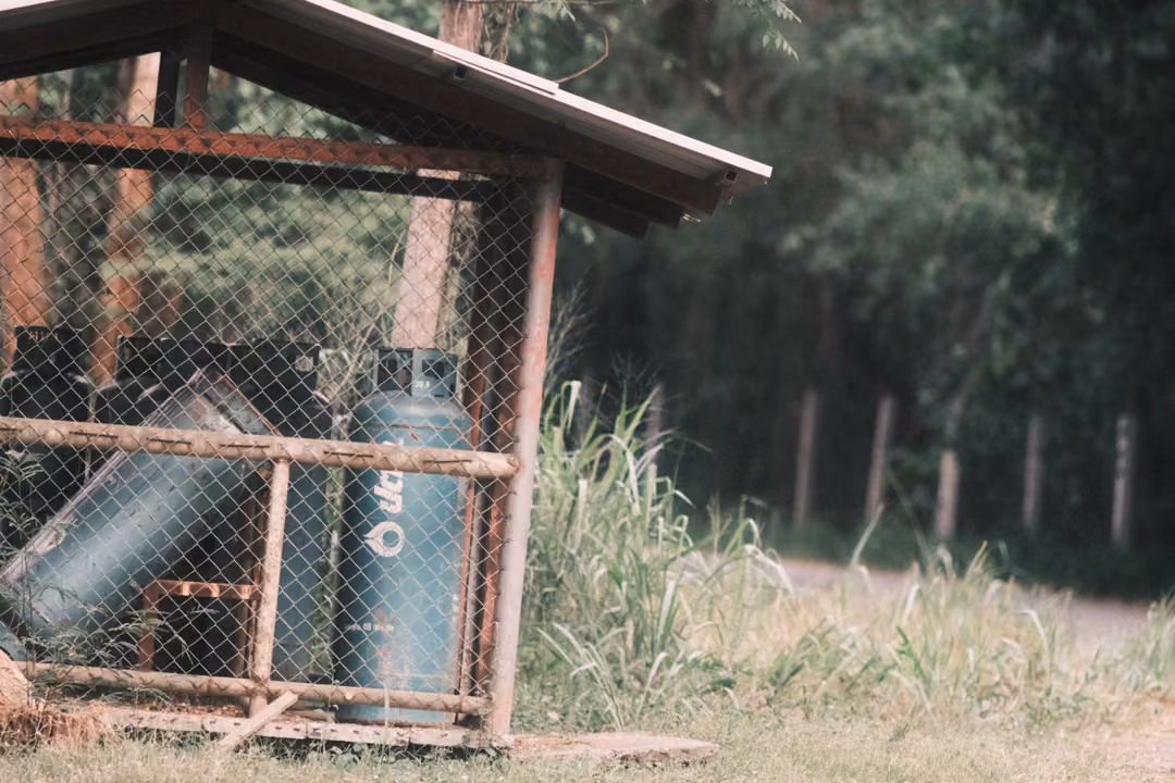 Wire cage with green gas cylinders under a slanted roof, tall grass and a post fence to the right