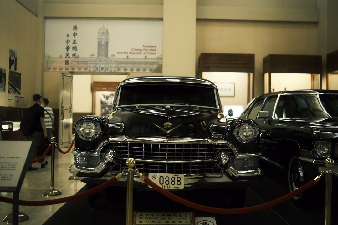 Front of a black vintage car with plate 0888 behind red ropes, a framed historical poster on the wall above