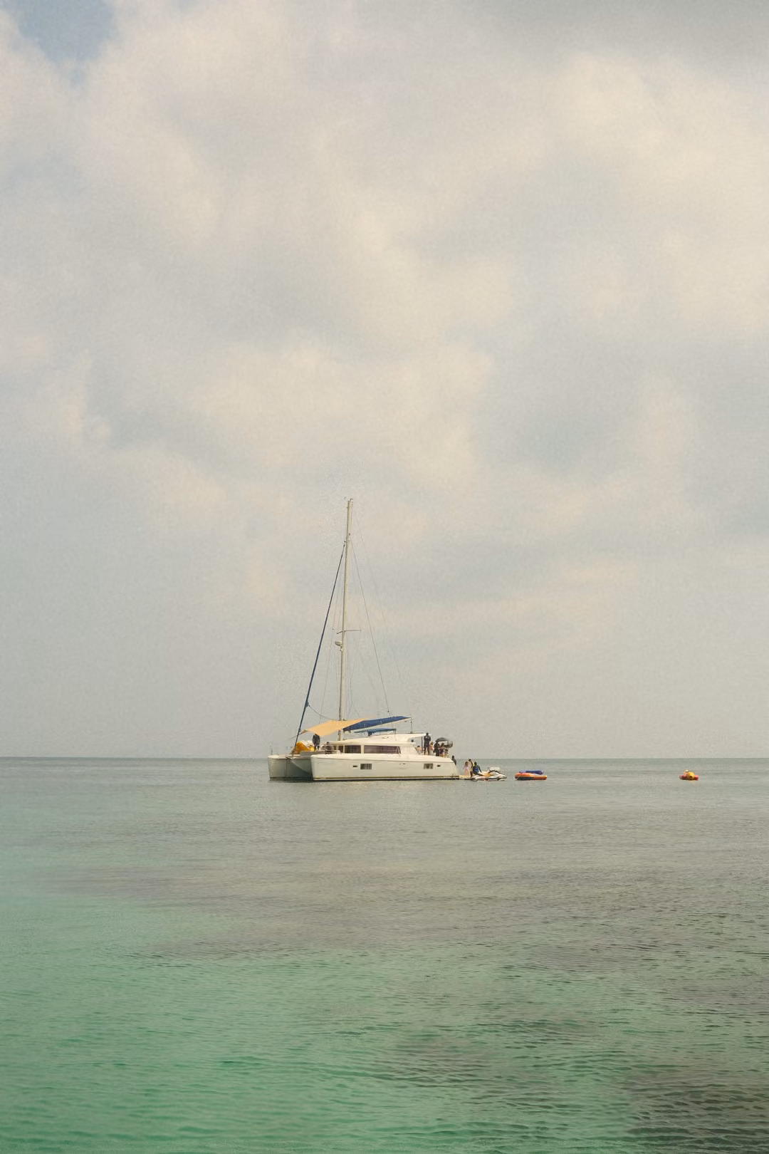 A catamaran sailboat anchored on clear water, with small floats and a dinghy beside it.