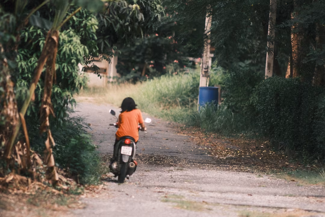 A rider in an orange shirt on a scooter heading away down a tree-lined lane past a blue barrel.
