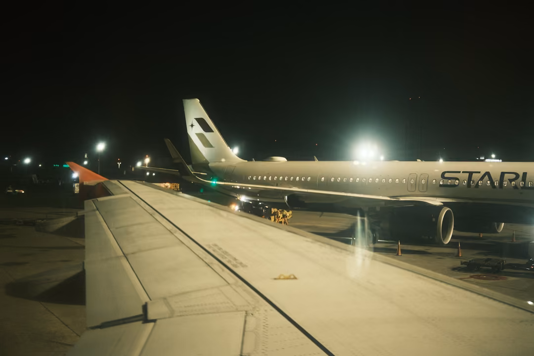 Night view over an aircraft wing toward another airliner on a lit apron, lettering 'STAR' on its fuselage
