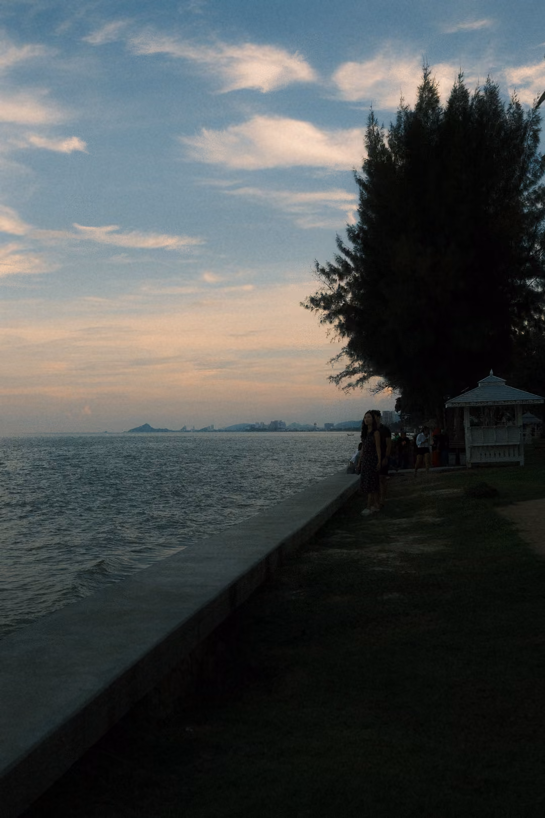 Seawall path along a calm sea at dusk, a dark pine and small white gazebo on the right, distant figures