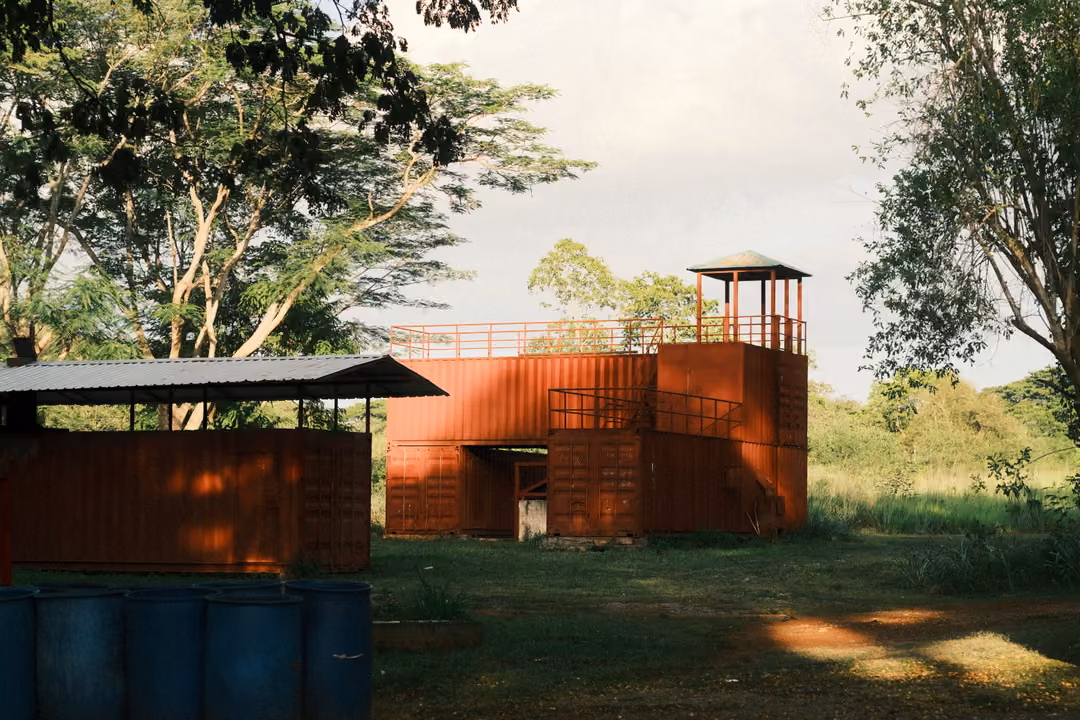 Rust-red container buildings with a small lookout tower and blue barrels under a shelter, trees and pale sky behind.