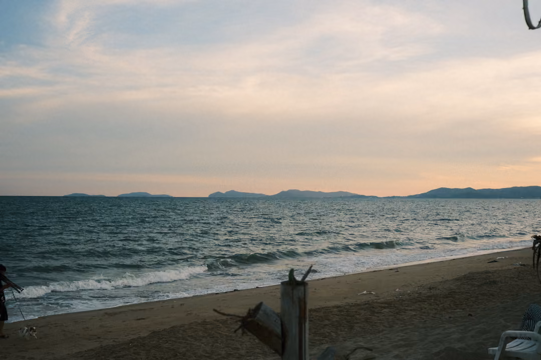 Sandy beach at sunset with gentle waves, distant hills on the horizon and a broken wooden post in front
