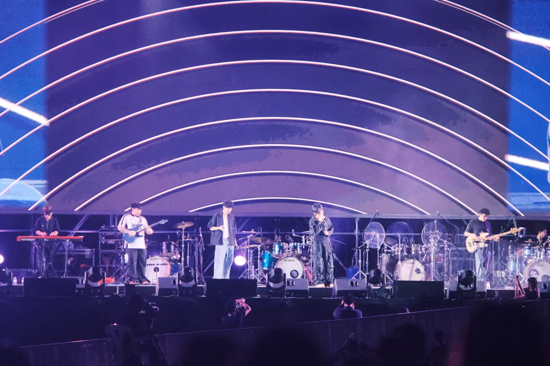 Stage with a purple arc-patterned screen, the band lined up across the platform under white streaks of light.