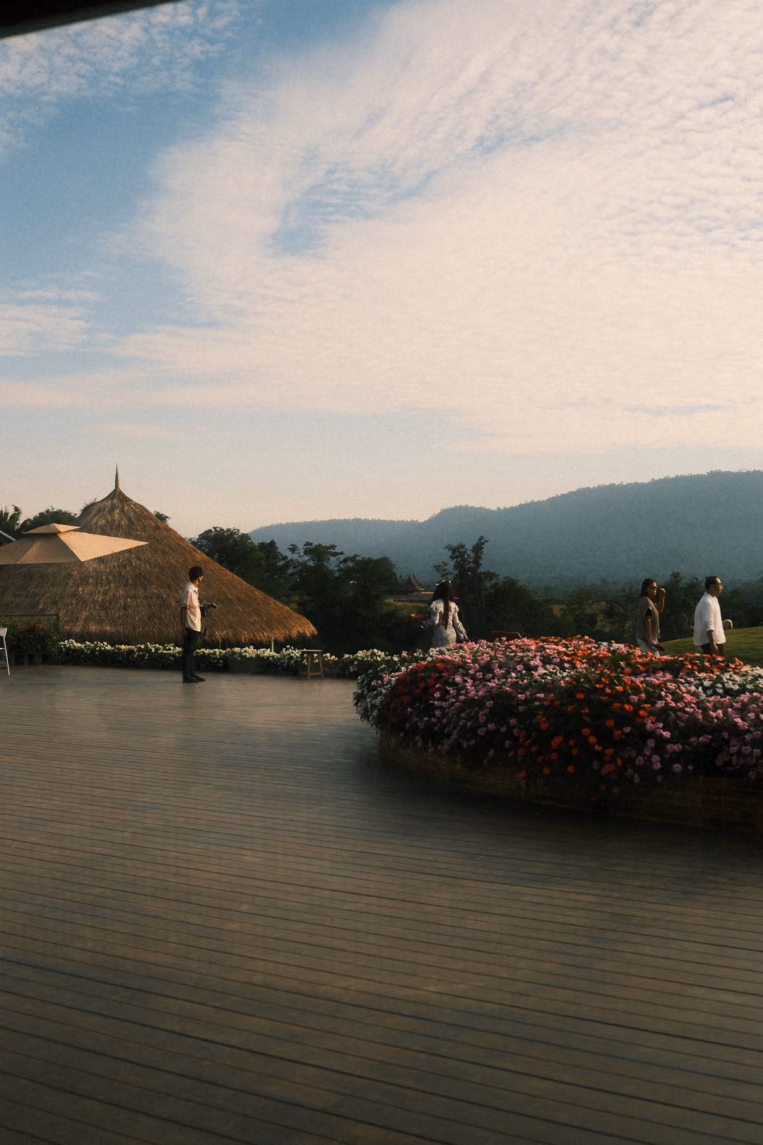 A wooden deck with a round bed of red and pink flowers, a thatched roof and mountains under blue sky.
