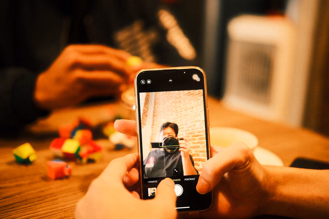 Hands holding a phone showing a camera screen pointed at a person with a camera, colourful toy pieces on a wooden table