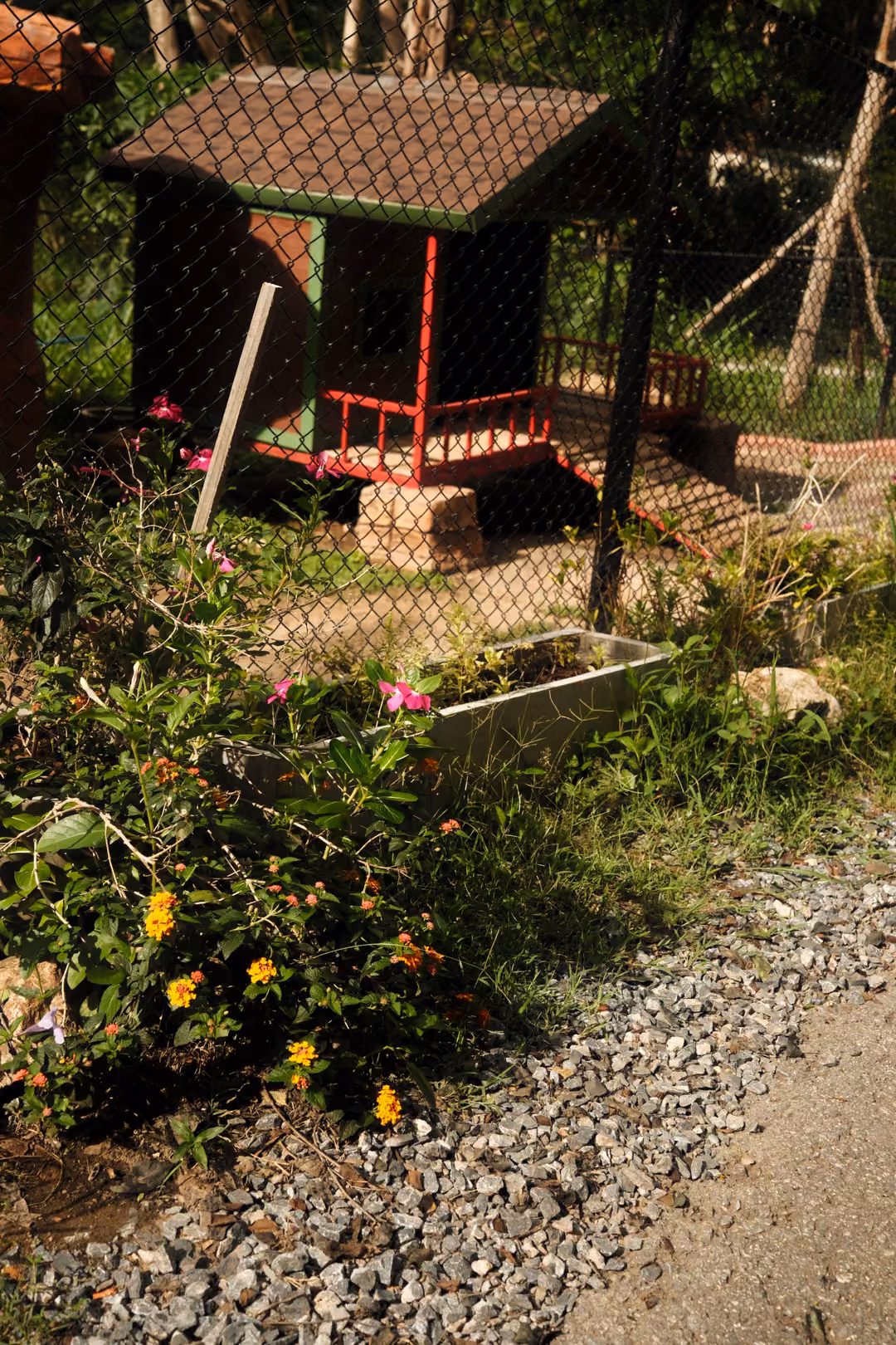 Small hut with a brown roof and red rails behind a chain-link fence, with flowers and gravel in front
