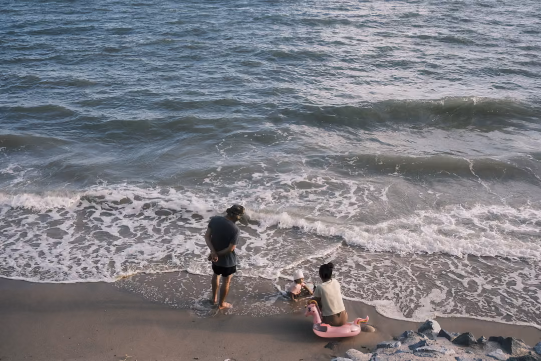 Seen from above, an adult stands in foamy surf beside a child sitting on the sand with a pink float