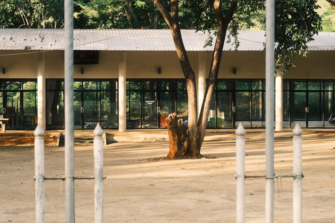 A long single-storey building with glass doors and a metal roof, white poles and a tree stump in the dirt yard.