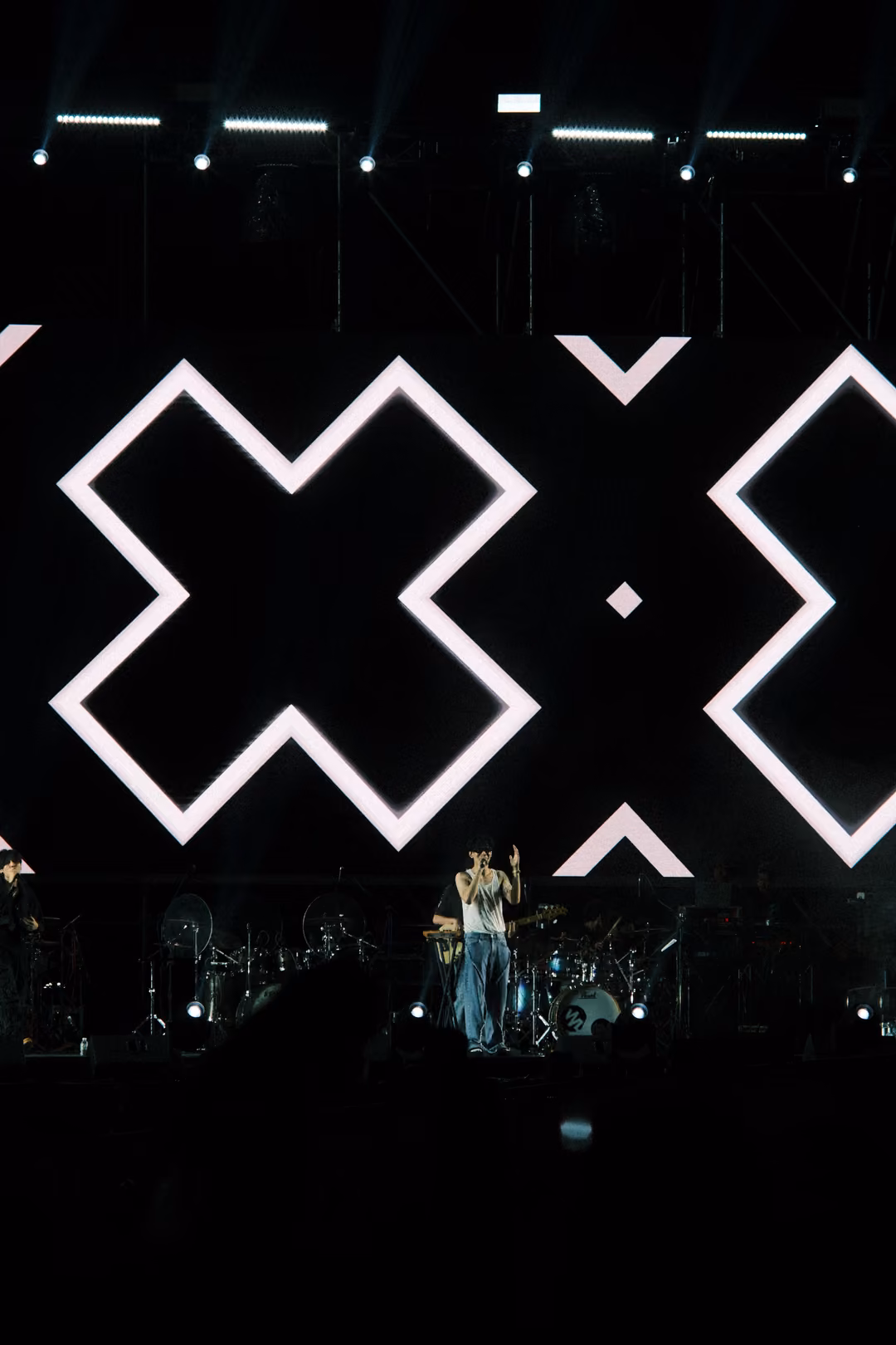 Vertical view of a screen with large white X outlines over black, a single performer standing on stage below.