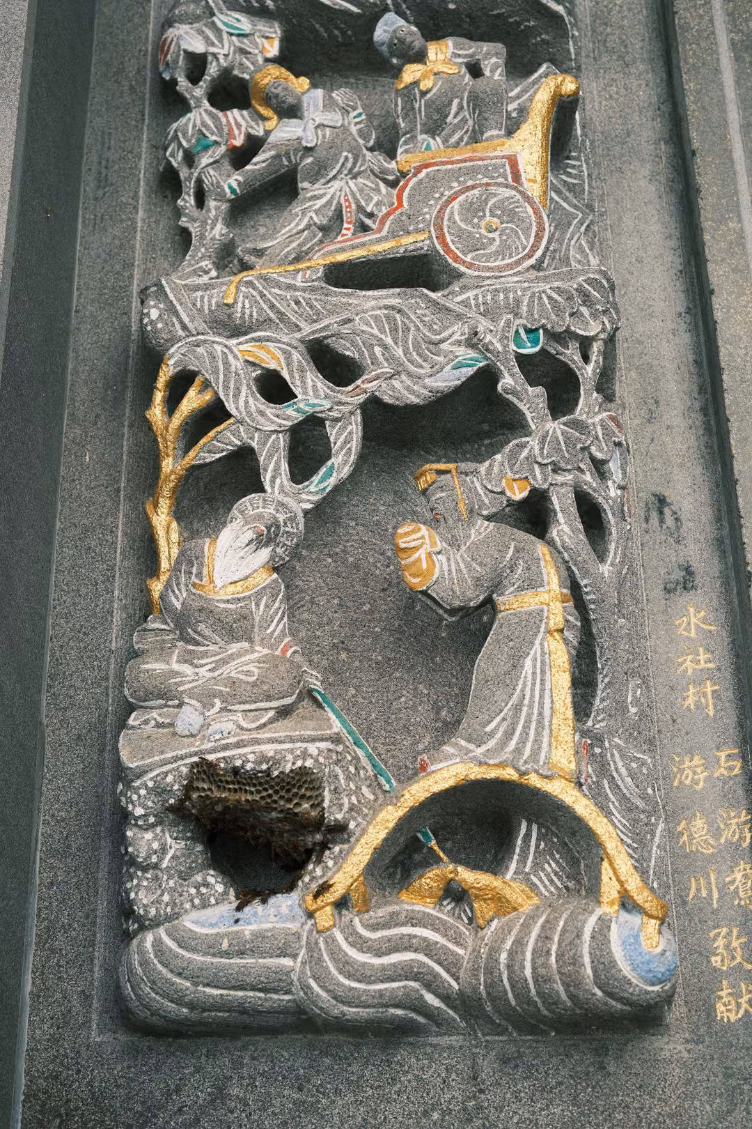 Carved stone panel of figures, a chariot and trees with gold detail, a small nest lodged in the lower carving