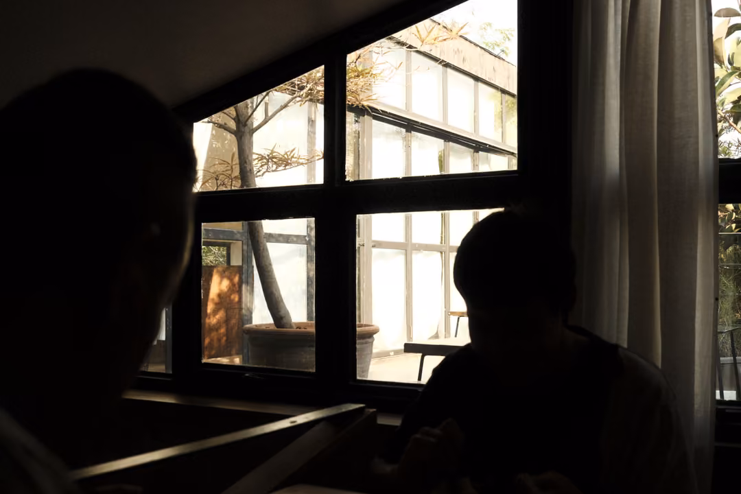 Two silhouetted people seated by tall black-framed windows looking out onto bright trees