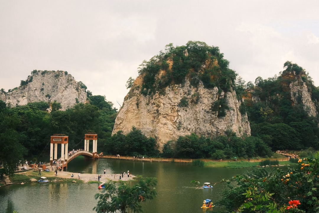 Wide view of a lake with a bridge, people on the shore, pedal boats, and three tree-capped limestone crags