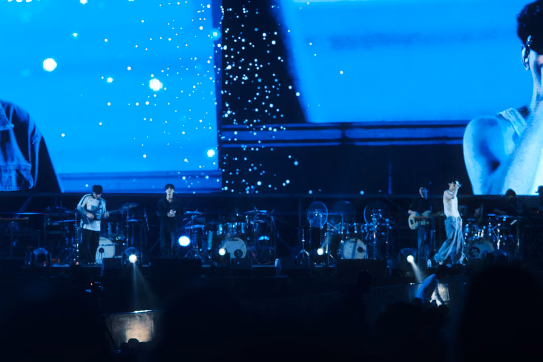 Stage screen split into panels of falling white specks and a cropped shoulder, band performing in blue light below.