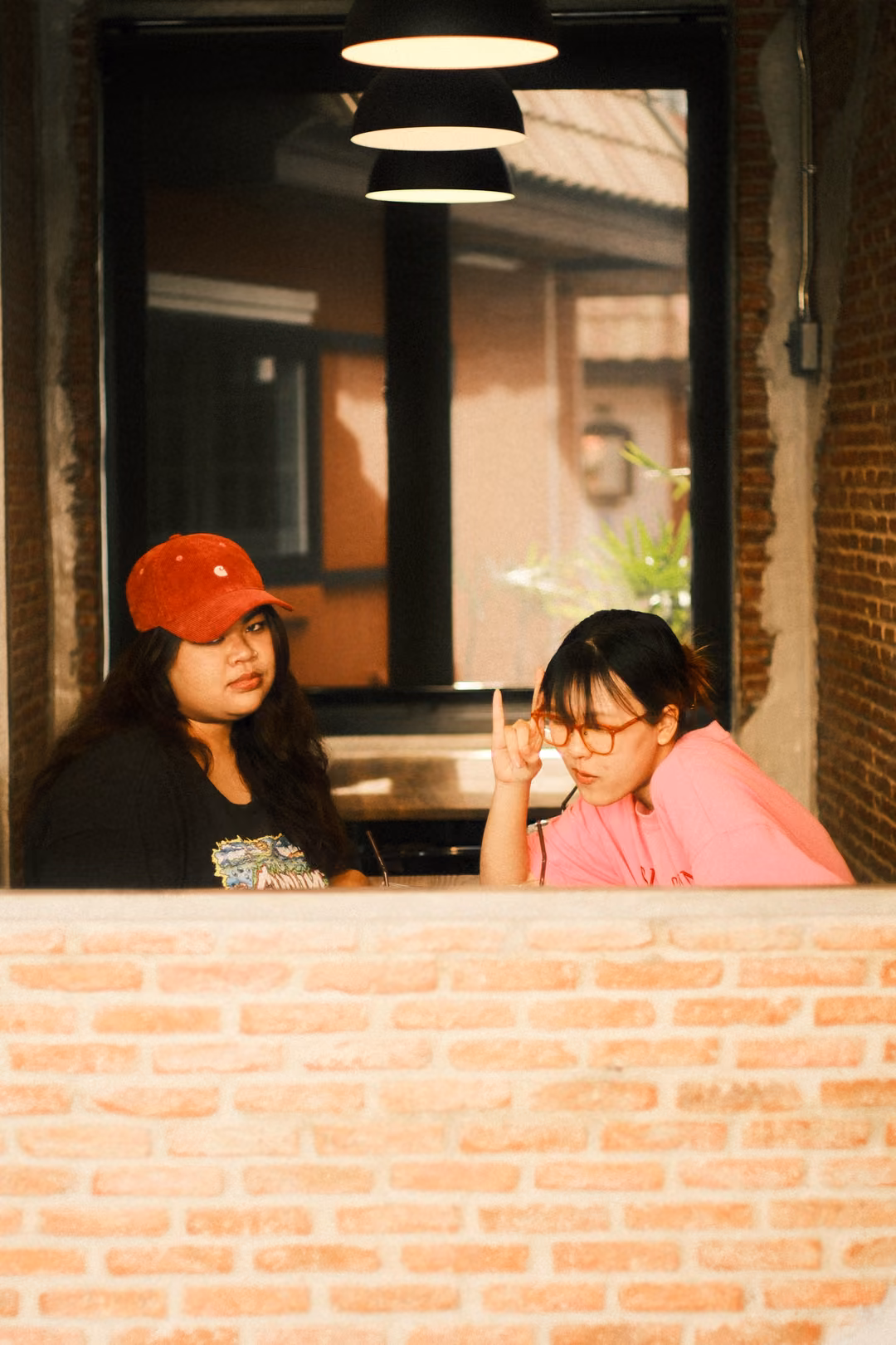 Two women at a window, the one in pink leaning in with one finger raised, the other in a red cap looking down