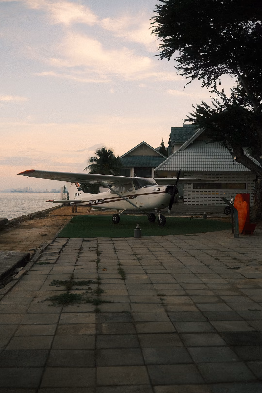 Small white propeller plane parked on a lawn beside a paved seafront walkway at dusk