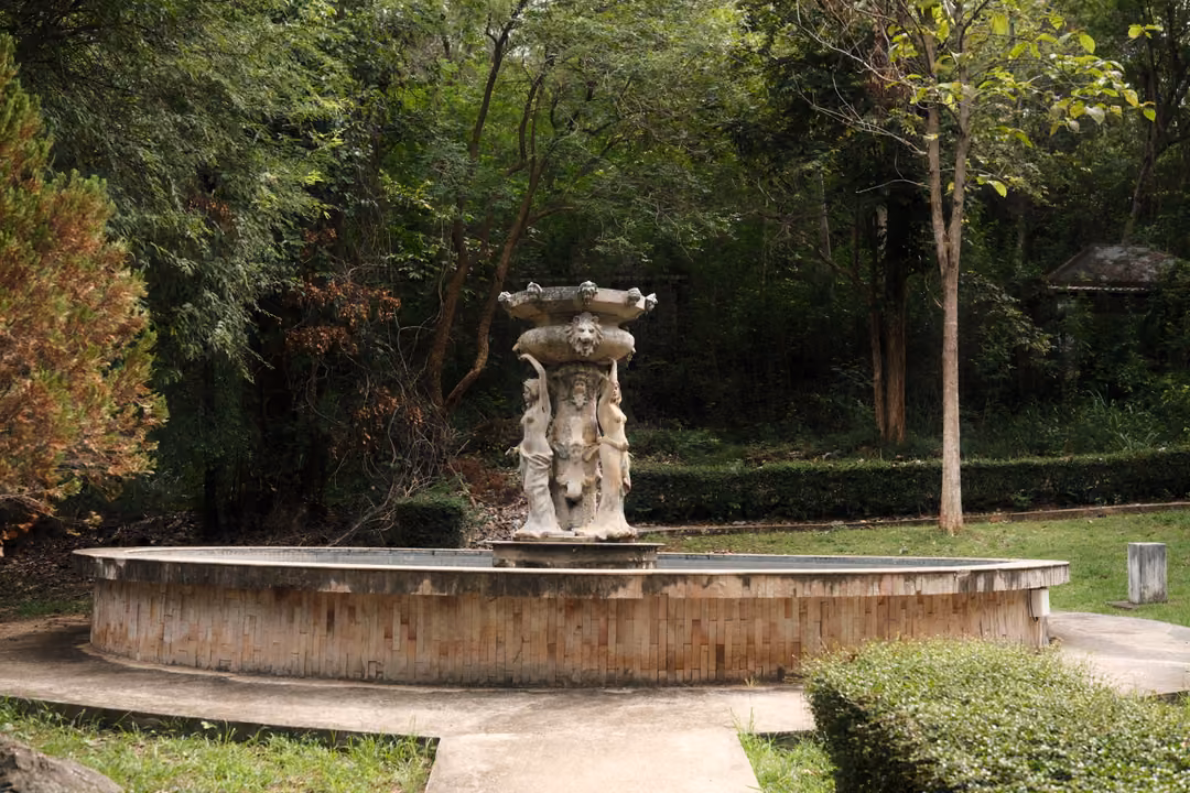 Ornate stone fountain in a round basin surrounded by lawn, clipped hedges and tall trees