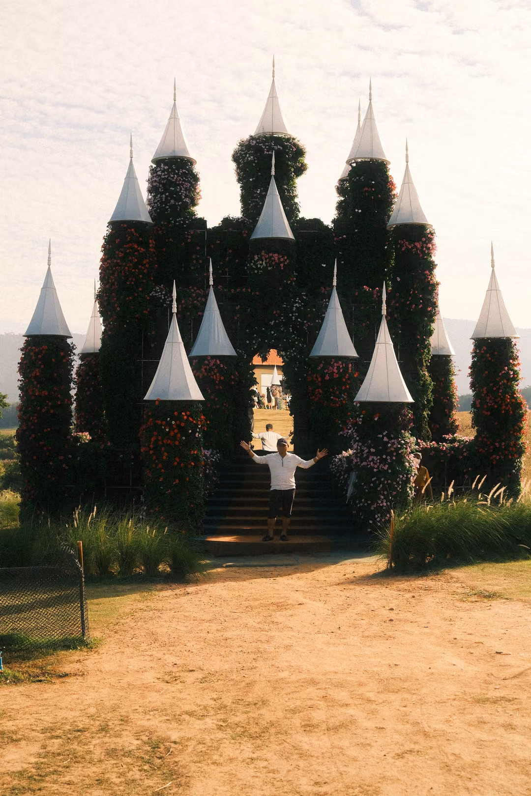 A person stands with arms outstretched on the steps of the flower-covered castle, spires above.