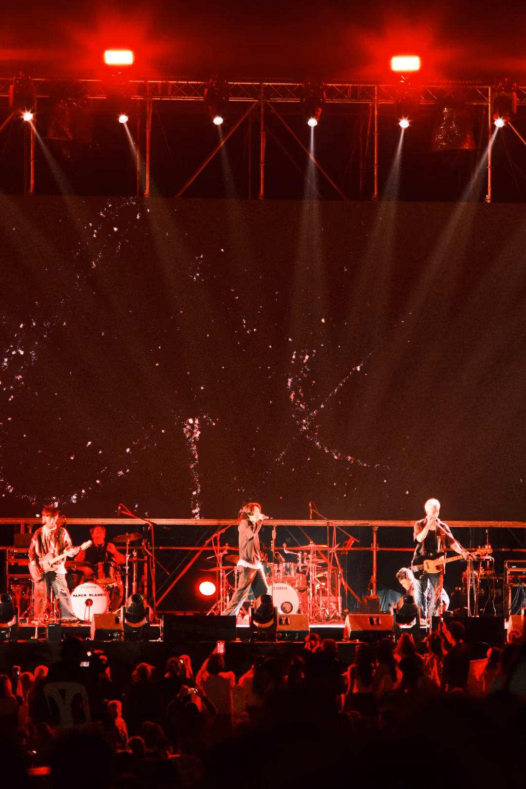Vertical view of a red-lit stage with spotlights fanning down, three musicians performing, crowd below.