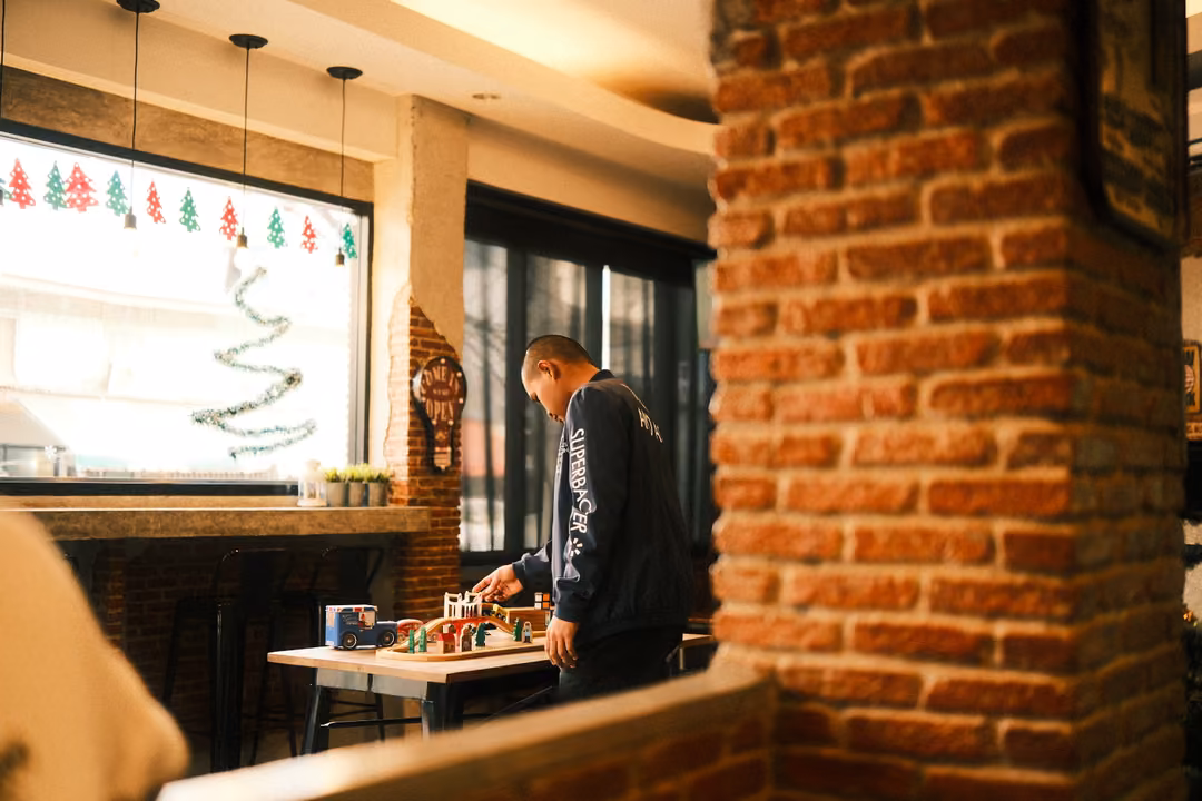 Café interior with a brick pillar, a person bending over a table of small toys near a window with Christmas decorations