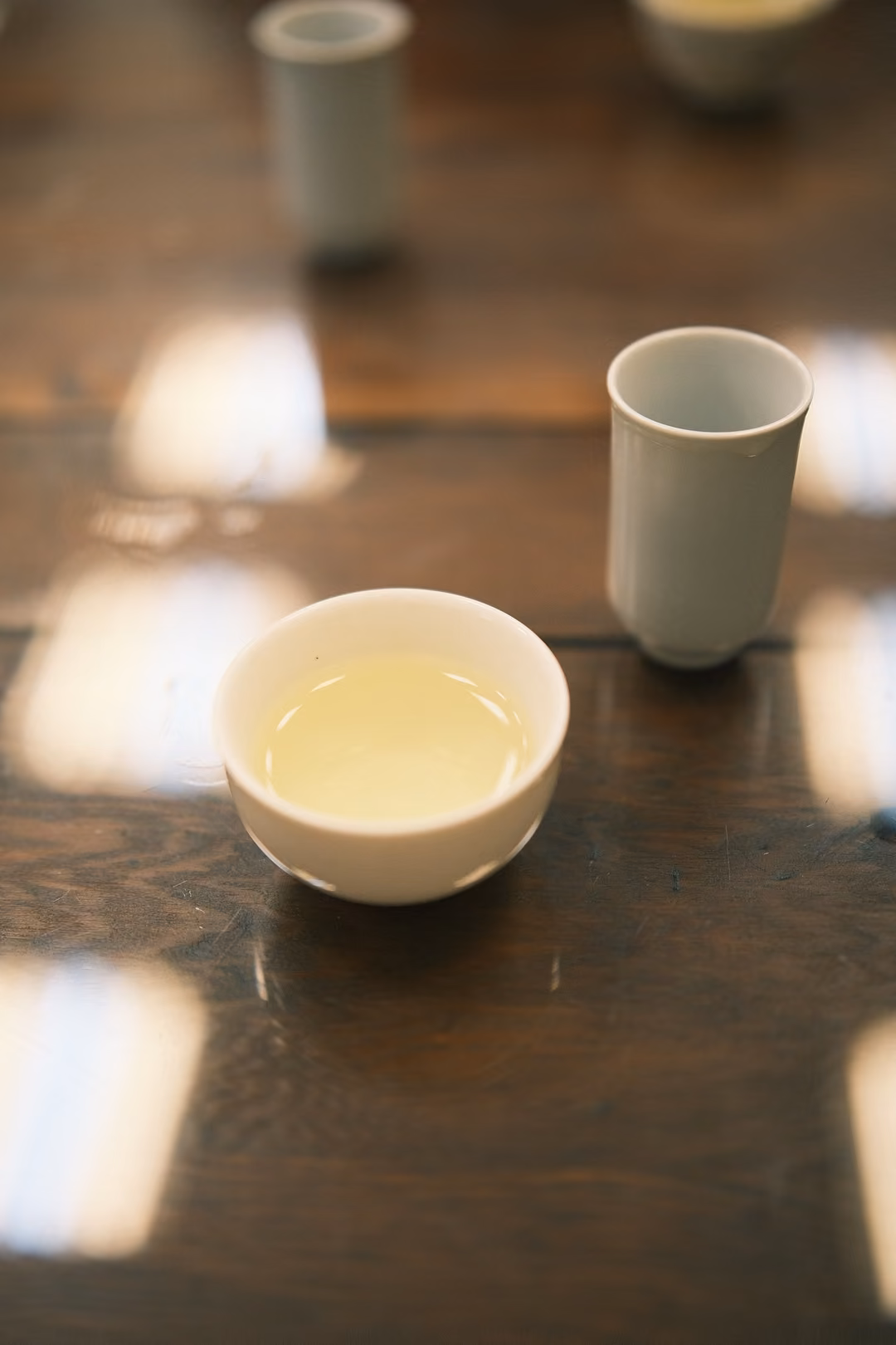 Small white bowl of pale yellow tea on a dark glossy table, a white tasting cup behind it