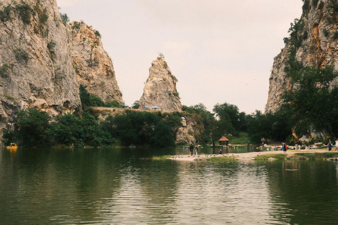 Wide green lake with a pointed rock spire between cliffs, visitors gathered along a distant shoreline